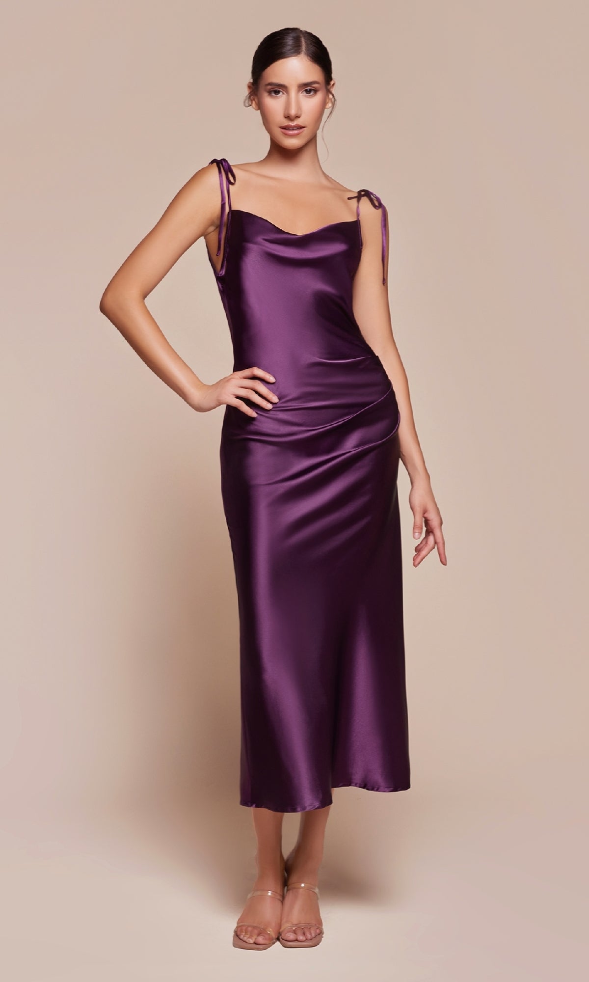 A woman stands against a beige background wearing the Midi Wedding-Guest Slip Dress BD103 in deep purple satin with shoulder ties, paired with clear strap heels.